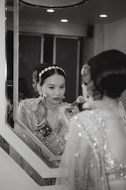 A woman in traditional attire is applying makeup in front of a mirror. She wears a heavily embellished outfit and jewelry, including bracelets and a headband with gem-like decorations. The setting appears elegant and is softly lit.