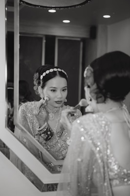 A woman in traditional attire is applying makeup in front of a mirror. She wears a heavily embellished outfit and jewelry, including bracelets and a headband with gem-like decorations. The setting appears elegant and is softly lit.