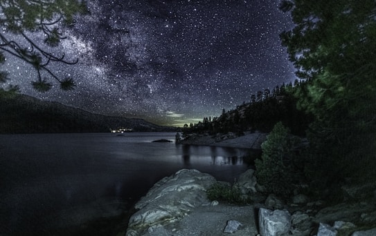 A breathtaking night sky filled with countless stars stretches over a serene lake surrounded by rocky cliffs and dense pine trees. The reflection of the starlit sky shimmers on the calm water's surface, creating a mirror-like effect. The landscape is bathed in a soft, dark blue hue, with the silhouette of distant mountains adding depth to the scene.