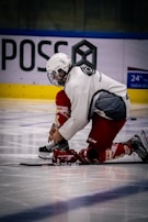 A hockey player dressed in red and white kneels on the ice while tying the laces of a skate. The player is wearing a helmet with a face cage and appears focused on the task. In the background, there is some advertising and a blue and yellow barrier.