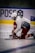A hockey player dressed in red and white kneels on the ice while tying the laces of a skate. The player is wearing a helmet with a face cage and appears focused on the task. In the background, there is some advertising and a blue and yellow barrier.