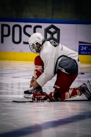 A hockey player dressed in red and white kneels on the ice while tying the laces of a skate. The player is wearing a helmet with a face cage and appears focused on the task. In the background, there is some advertising and a blue and yellow barrier.