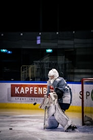 A hockey goalie wearing a dark jersey and white protective gear stands in front of the goal. The ice rink features advertisements on the barriers, and the lighting suggests an indoor arena.