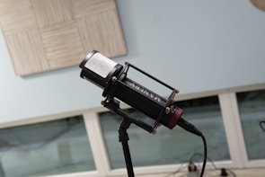 A professional microphone is set up on a stand in front of a window with a blurred outdoor view. The microphone is black with a cylindrical shape and has some metallic screws and components. Behind the microphone, part of an acoustic panel is visible on the wall, suggesting a recording or studio environment. The environment appears clean and well-organized.
