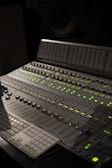 A professional audio mixing console with numerous sliders, knobs, and digital displays illuminated by green lights. The control panel is advanced with multiple sections for precise audio adjustments, and a keyboard is partially visible.