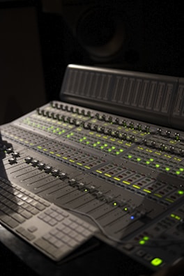 A professional audio mixing console with numerous sliders, knobs, and digital displays illuminated by green lights. The control panel is advanced with multiple sections for precise audio adjustments, and a keyboard is partially visible.