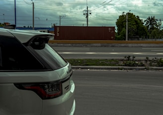 A white SUV parked on the side of a road with a container truck moving in the background. The sky is cloudy, and there are power lines above and trees visible in the distance.