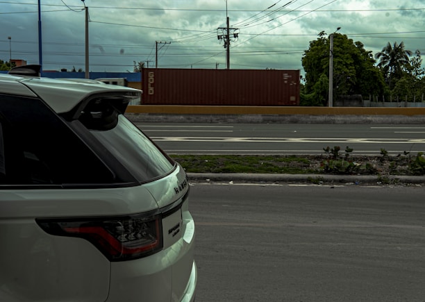A white SUV parked on the side of a road with a container truck moving in the background. The sky is cloudy, and there are power lines above and trees visible in the distance.