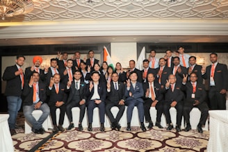A group of people dressed in formal attire with orange ties are posing for a group photo indoors. The background features intricate ceiling patterns and two flags, one of which is the Indian flag. The individuals are showing gestures with their hands, and there is a sense of unity and formality.