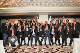 A group of people dressed in formal attire with orange ties are posing for a group photo indoors. The background features intricate ceiling patterns and two flags, one of which is the Indian flag. The individuals are showing gestures with their hands, and there is a sense of unity and formality.