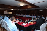 A large group of people seated around a long, rectangular conference table in a well-lit room with wooden paneling and chandeliers. There are water bottles, glasses, notebooks, and other meeting materials on the table. A couple of whiteboards are visible in the background, and the participants are mostly wearing similar attire with name tags.