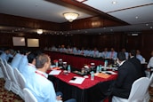 A large group of people seated around a long, rectangular conference table in a well-lit room with wooden paneling and chandeliers. There are water bottles, glasses, notebooks, and other meeting materials on the table. A couple of whiteboards are visible in the background, and the participants are mostly wearing similar attire with name tags.