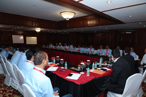 A large group of people seated around a long, rectangular conference table in a well-lit room with wooden paneling and chandeliers. There are water bottles, glasses, notebooks, and other meeting materials on the table. A couple of whiteboards are visible in the background, and the participants are mostly wearing similar attire with name tags.