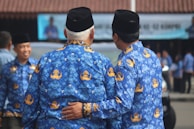 Two men wearing matching blue patterned batik shirts and black hats are standing side by side, with one man's arm around the other's back. They appear to be in a public setting with blurred figures and a building in the background.