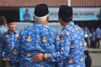 Two men wearing matching blue patterned batik shirts and black hats are standing side by side, with one man's arm around the other's back. They appear to be in a public setting with blurred figures and a building in the background.