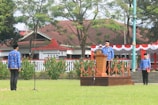 A group of people wearing traditional blue uniforms stands outdoors. One person is speaking at a wooden podium adorned with decorative red and white fabric, suggesting a formal or ceremonial event. The setting includes a grassy area with neatly arranged bushes and trees. In the background, there is a building with a brown roof, partially obscured by trees.