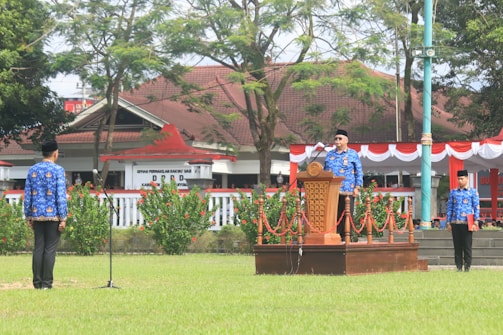 A group of people wearing traditional blue uniforms stands outdoors. One person is speaking at a wooden podium adorned with decorative red and white fabric, suggesting a formal or ceremonial event. The setting includes a grassy area with neatly arranged bushes and trees. In the background, there is a building with a brown roof, partially obscured by trees.