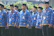 A group of men are standing in formation, wearing matching blue batik shirts and black hats. They each have identification badges hanging around their necks and appear to be part of an organized event or ceremony. The setting includes green foliage in the background.
