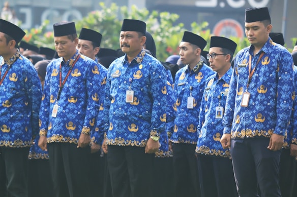 A group of men are standing in formation, wearing matching blue batik shirts and black hats. They each have identification badges hanging around their necks and appear to be part of an organized event or ceremony. The setting includes green foliage in the background.