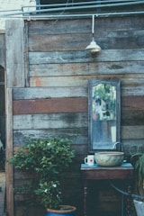 An outdoor wooden wall with a weathered appearance has a large mirror mounted on it. Beneath the mirror, there is a small wooden table holding a decorative bowl, a mug with a heart design, and a faucet attached to a hose. A small potted plant with green leaves and small white flowers is placed below the table. A vintage-style lantern is mounted above the mirror, hanging from an electric wire running across the top of the wall.