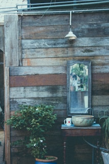 An outdoor wooden wall with a weathered appearance has a large mirror mounted on it. Beneath the mirror, there is a small wooden table holding a decorative bowl, a mug with a heart design, and a faucet attached to a hose. A small potted plant with green leaves and small white flowers is placed below the table. A vintage-style lantern is mounted above the mirror, hanging from an electric wire running across the top of the wall.