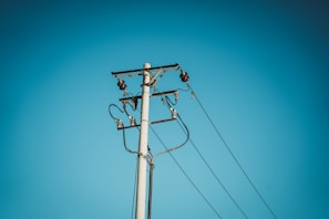 A utility pole with several electrical wires and insulators stretches up into a clear blue sky. The pole is tall, painted white or light grey, and it is equipped with multiple crossarms and metal components.