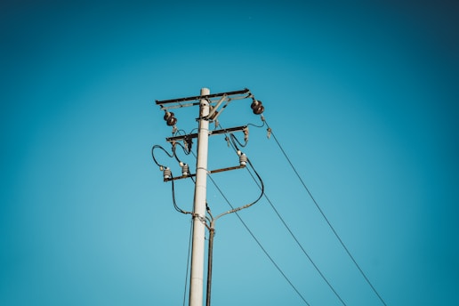 A utility pole with several electrical wires and insulators stretches up into a clear blue sky. The pole is tall, painted white or light grey, and it is equipped with multiple crossarms and metal components.