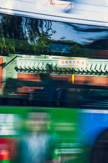 A blurred image of a moving bus passing by a traditional Chinese architectural structure. The background reveals a sign with Chinese characters and the words 'Gulou Dongdajie'. The reflection of trees and the presence of vibrant colors create a sense of motion.