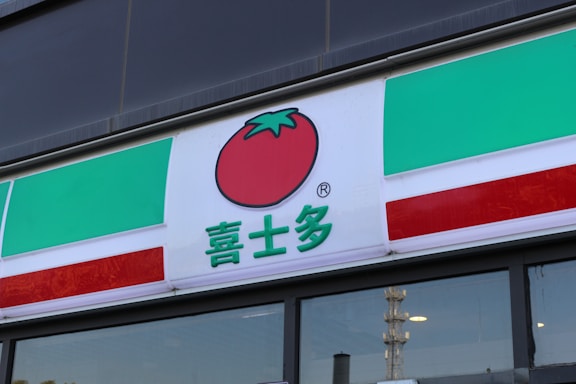 A storefront sign with a tomato icon above Chinese characters. The sign has green, white, and red horizontal stripes, resembling the colors of the Italian flag. The background above the sign is part of the building's facade, possibly made of glass or a dark material.