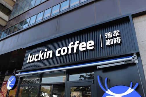 A modern storefront features the sign 'luckin coffee' in bold, white letters on a black background. The sign also includes Chinese characters next to it. The facade of the building is sleek and urban with floor-to-ceiling windows on the upper floors.