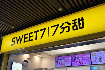A bright yellow sign with bold black text featuring the words 'SWEET7' and Chinese characters. Below the sign, digital screens display colorful images of beverages and desserts, with one screen showing a polar bear alongside drink options. The store is well-lit with a modern interior, featuring ceiling lights and a small air conditioning unit.
