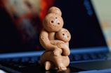 A wooden sculpture depicting a mother embracing a child, both with smiling expressions. The sculpture is positioned on the keyboard of an open laptop, with a blurred background featuring warm colors.