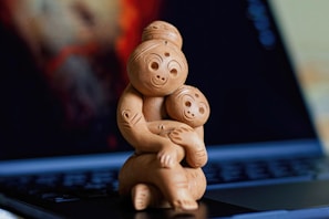 A wooden sculpture depicting a mother embracing a child, both with smiling expressions. The sculpture is positioned on the keyboard of an open laptop, with a blurred background featuring warm colors.