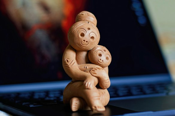 A wooden sculpture depicting a mother embracing a child, both with smiling expressions. The sculpture is positioned on the keyboard of an open laptop, with a blurred background featuring warm colors.