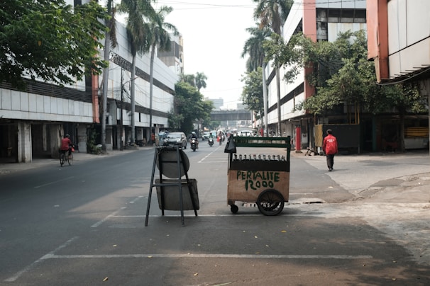 An urban street with several people and vehicles. A small cart labeled 'PERTALITE 12000' is positioned in the foreground, slightly to the right. The street is flanked by tall buildings and lined with trees. A person in a red jacket walks near the cart, while another is cycling on the left side of the street. Several other vehicles, including motorcycles, are visible further down the road.