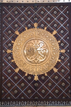 An ornate door features a prominent circular design in the center, with intricate Arabic calligraphy surrounded by geometric patterns. The wood is rich and dark, with metal accents and a golden-hued centerpiece showcasing elaborate detailing.