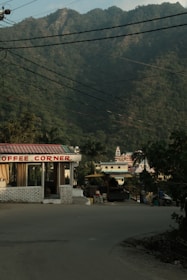 A roadside coffee shop with a red and white sign reading 'Coffee Corner' is situated beside a street. In the background, lush green mountains rise prominently, and there are several buildings including a structure that resembles a temple. Overhead, numerous power lines intersect the view.