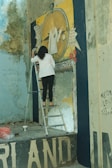 A person standing on a metal ladder, painting a mural on a wall. The mural depicts a large compass with text and a cloak draped over it. The room appears worn, with peeling paint and visible graffiti. Paint cans and a plastic bag are on the floor.