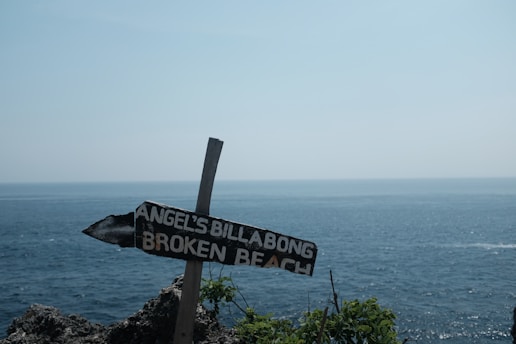 A weathered wooden sign pointing towards 'Angel's Billabong' and 'Broken Beach' stands against a backdrop of a vast ocean. The sign is planted in rocky terrain with sparse vegetation, and the scene is illuminated by a clear, bright sky.
