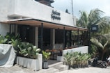 A modern cafe with a minimalist design features an open patio with wooden tables and chairs. Lush green plants line the entrance, providing a vibrant contrast to the light grey concrete walls. A sign above the entrance reads 'Asian Spoon.' The surroundings include palm trees and parked scooters, suggesting a tropical setting.