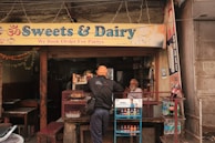 A small shop with a sign reading 'Sweets & Dairy' offers various beverages and snacks. A person with a turban is seen interacting with the shopkeeper, who is seated behind the counter. The shop is adorned with colorful decorations and is set in an urban environment.