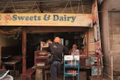 A small shop with a sign reading 'Sweets & Dairy' offers various beverages and snacks. A person with a turban is seen interacting with the shopkeeper, who is seated behind the counter. The shop is adorned with colorful decorations and is set in an urban environment.