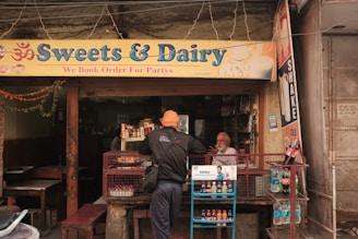 A small shop with a sign reading 'Sweets & Dairy' offers various beverages and snacks. A person with a turban is seen interacting with the shopkeeper, who is seated behind the counter. The shop is adorned with colorful decorations and is set in an urban environment.