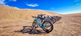 A line of mountain bikes is parked on a sandy terrain with a large sand dune in the background. The sky above is clear and bright blue, suggesting a sunny day. A person stands behind the bikes with arms outstretched, adding a sense of scale to the scene.