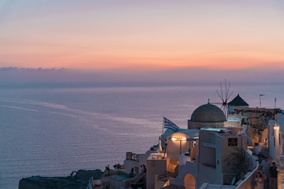 A serene view of a coastal village with white-washed buildings, a traditional windmill, and a Greek flag, overlooking the calm sea at sunset. The sky is painted with soft hues of pink and orange, casting a warm glow over the scene.