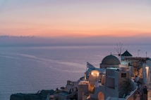 A serene view of a coastal village with white-washed buildings, a traditional windmill, and a Greek flag, overlooking the calm sea at sunset. The sky is painted with soft hues of pink and orange, casting a warm glow over the scene.