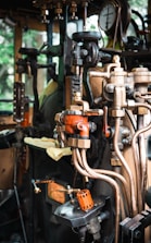 A detailed view of a complex array of industrial machinery components featuring valves, pipes, and gauges. The setup appears to be part of a steam engine or similar mechanical system. The machinery includes various metallic elements, with some parts painted orange for visibility and a pressure gauge prominently displayed.