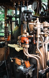 A detailed view of a complex array of industrial machinery components featuring valves, pipes, and gauges. The setup appears to be part of a steam engine or similar mechanical system. The machinery includes various metallic elements, with some parts painted orange for visibility and a pressure gauge prominently displayed.