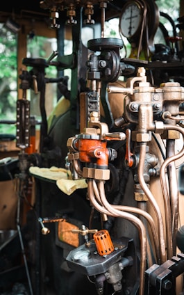 A detailed view of a complex array of industrial machinery components featuring valves, pipes, and gauges. The setup appears to be part of a steam engine or similar mechanical system. The machinery includes various metallic elements, with some parts painted orange for visibility and a pressure gauge prominently displayed.