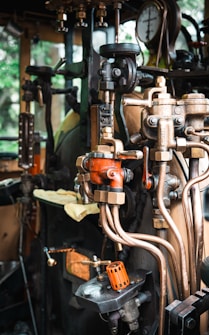A detailed view of a complex array of industrial machinery components featuring valves, pipes, and gauges. The setup appears to be part of a steam engine or similar mechanical system. The machinery includes various metallic elements, with some parts painted orange for visibility and a pressure gauge prominently displayed.